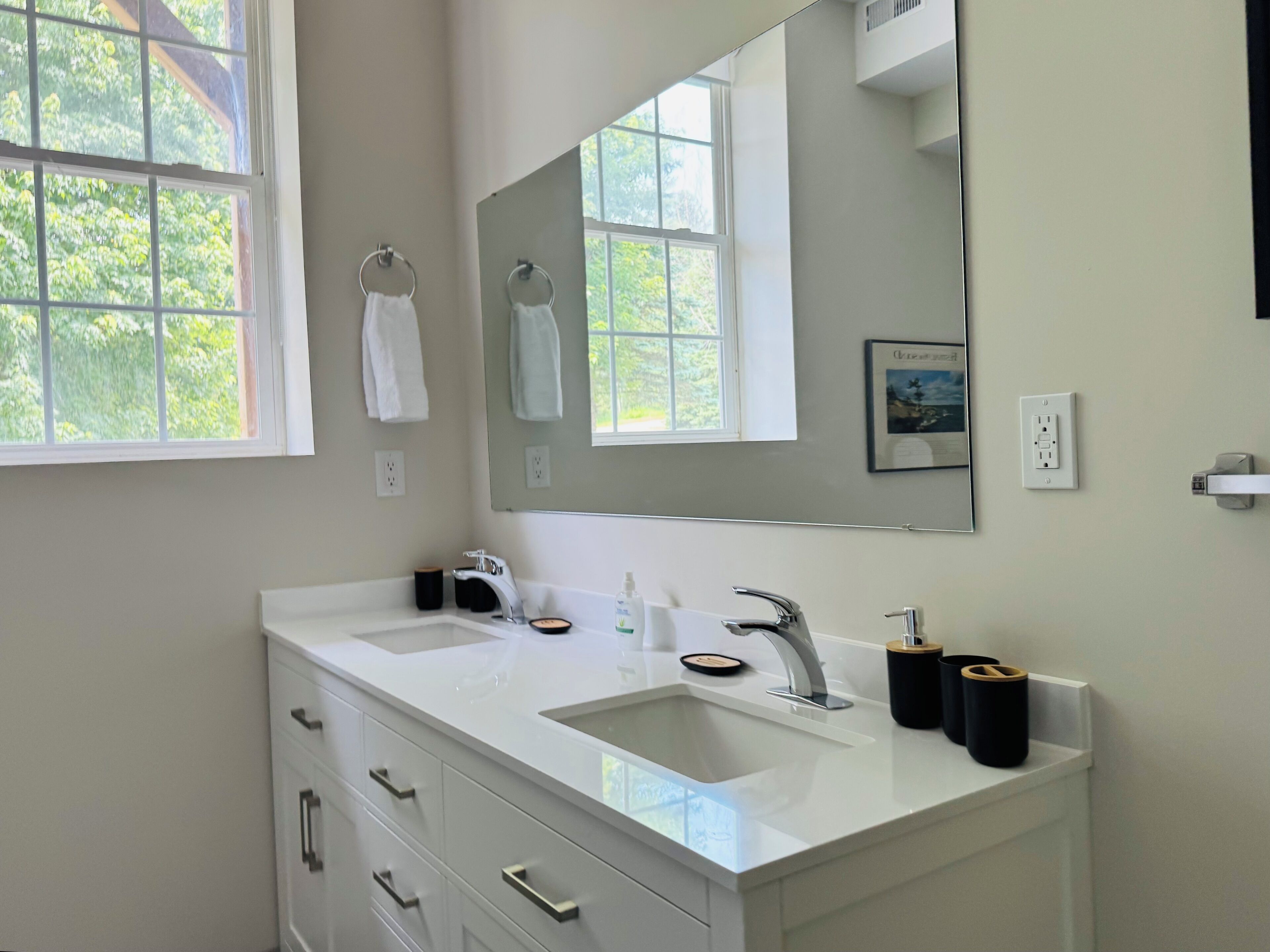 A modern bathroom with two sinks, a large mirror, and a window with a view of greenery.