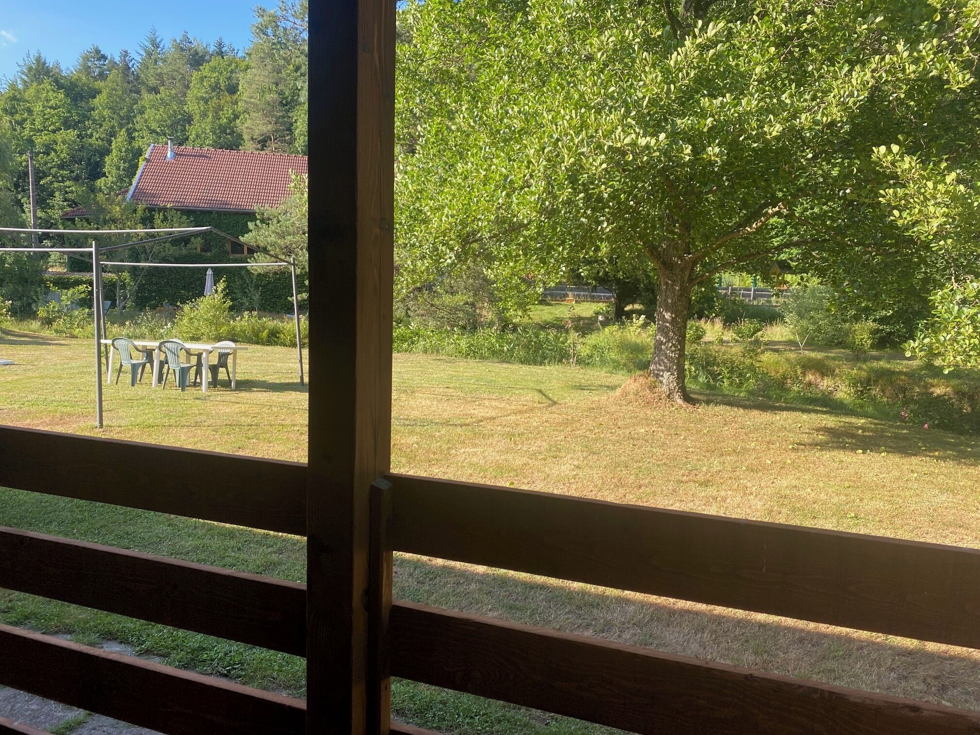 Vista a través de una ventana que muestra un área exterior con césped, una valla de madera, un árbol y un edificio con techo rojo al fondo.