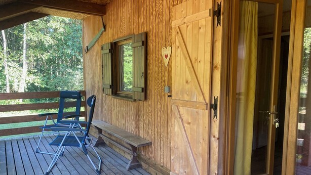 A wooden deck with a bench and a folding chair, a wooden door with a heart-shaped decoration, and a window with shutters. - The Kingfisher (Autrey)