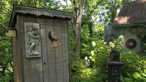 Un abri de jardin rustique en bois, orné d’un croissant de lune et d’un relief représentant une grenouille, entouré d’une végétation luxuriante et d’une petite lanterne.