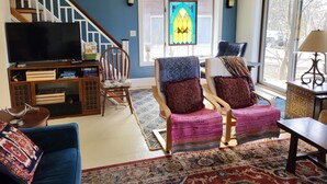 Un salon comprenant un téléviseur, un meuble de rangement en bois, deux fauteuils, un canapé, un tapis à motifs, un ventilateur de plafond et une fenêtre à vitraux.