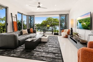 A modern living room with a sofa, coffee table, and TV, large windows with a view of greenery and a balcony.