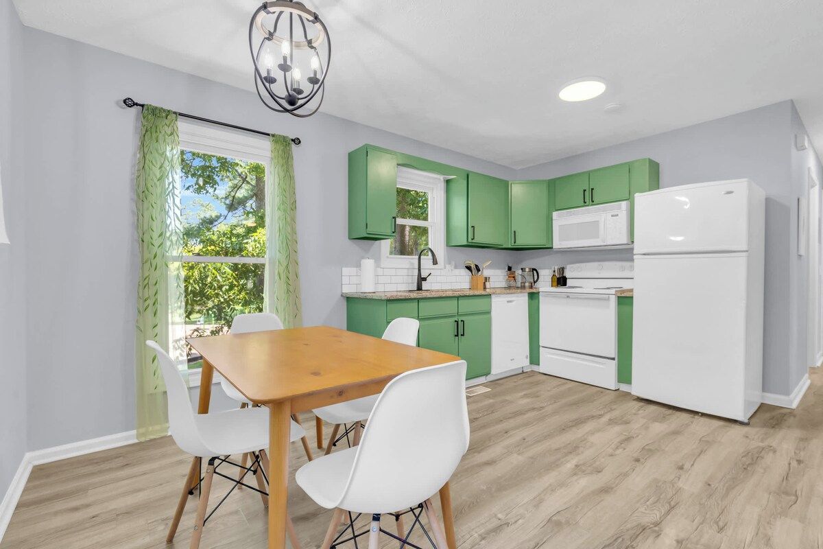 A modern kitchen with green cabinets, a white refrigerator, and a dining area with a wooden table and white chairs.