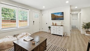 Amplia sala de estar con una mesa de centro de madera, un cómodo asiento tipo cojín, un mueble blanco y un televisor grande en la pared.