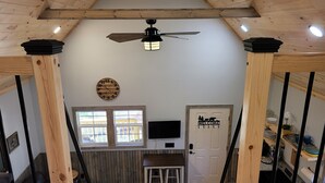 A room with a wooden ceiling, a ceiling fan, a clock, a welcome sign, a TV, a small table, and a window.