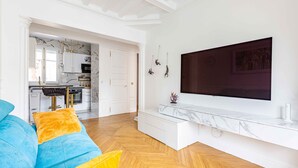 A modern living room with a blue sofa, a TV mounted on the wall, and a kitchen in the background.