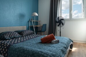 A bedroom with a bed, a desk, a chair, a standing fan, and a window with a view of the sky.