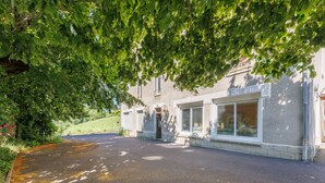 A building with a paved driveway leading to a gated entrance, surrounded by greenery and trees. - Source Fenestre - Proche Puy de Sancy (La Bourboule)