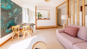 A modern living room with a dining area, a large floral painting, a wooden-framed mirror, and a soft pink sofa. - Source Fenestre - Proche Puy de Sancy (La Bourboule)