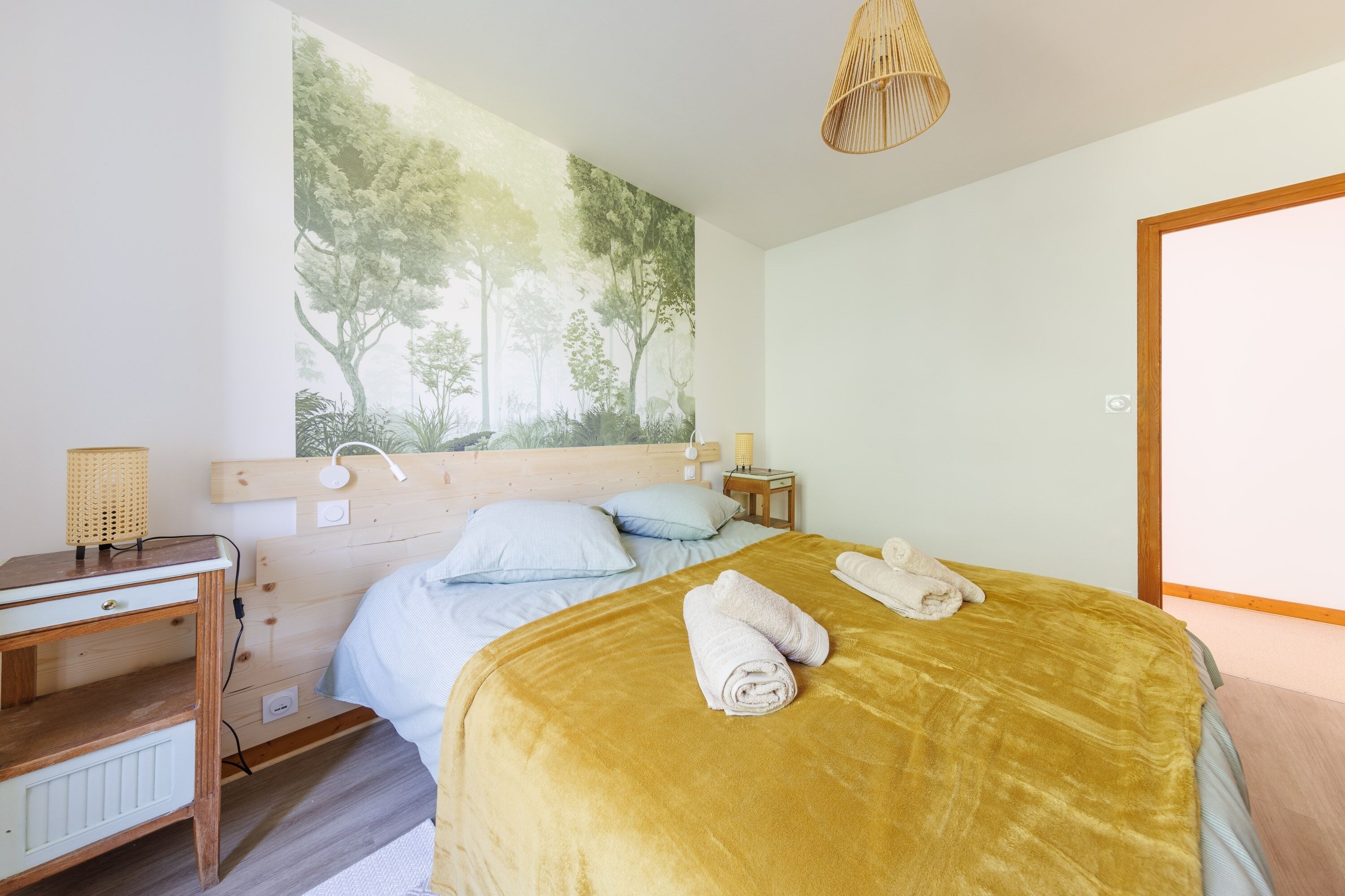 A bedroom with a bed, a nightstand, a desk, a lamp, and a wall mural.