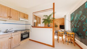 A compact kitchen with wooden cabinets, a built-in oven, and a dining area with a table and chairs. - Source Fenestre - Proche Puy de Sancy (La Bourboule)