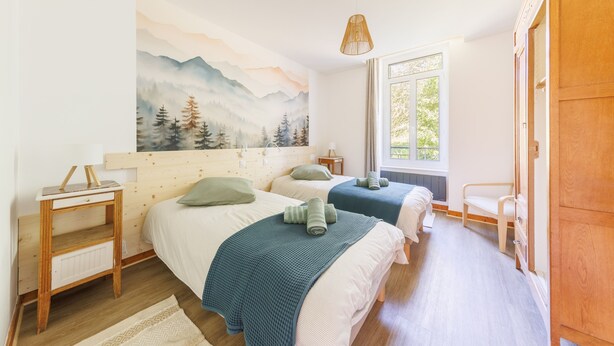 A bedroom with two beds, a wooden headboard, a painting of mountains, a window, and a wooden wardrobe. - Source Fenestre - Proche Puy de Sancy (La Bourboule)