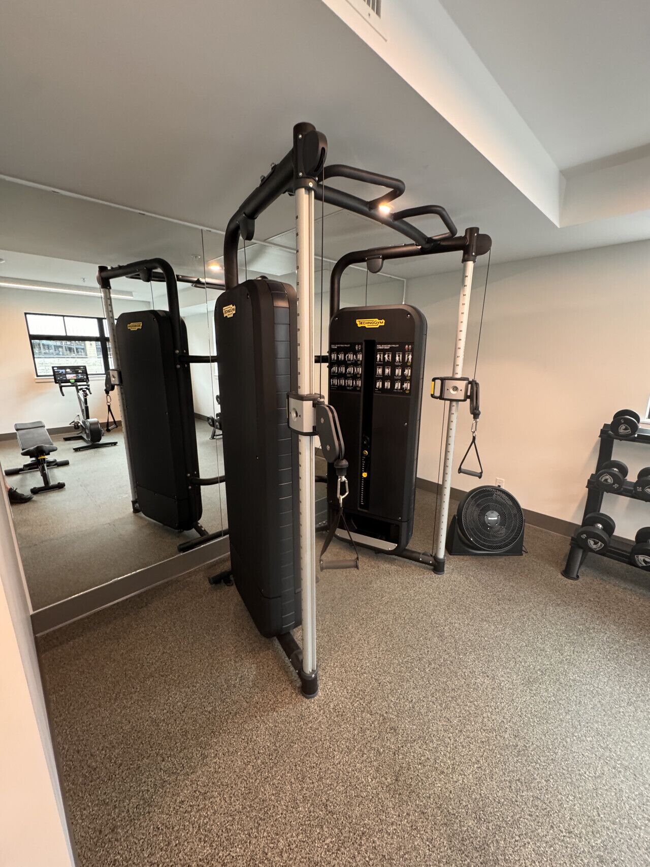 Un gimnasio con diverso equipo de ejercicios, incluyendo un rack para sentadillas, una banca de pesas y un ventilador.