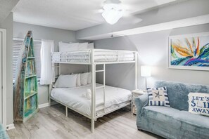 Un lit superposé avec une literie blanche, un canapé bleu avec des coussins à rayures, une lampe et une peinture abstraite colorée accrochée au mur.