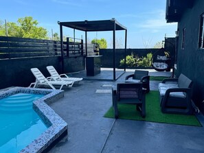 Un patio trasero con piscina, sillas de descanso, una zona de estar cubierta y un pequeño jardín.