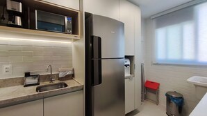 Une cuisine compacte équipée d’un four à micro-ondes encastré, d’un réfrigérateur en acier inoxydable, d’un évier et d’une fenêtre avec des stores.