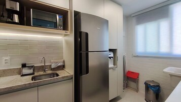 Uma cozinha compacta com micro-ondas integrado, frigorífico de aço inoxidável, uma pia e uma janela com persianas.