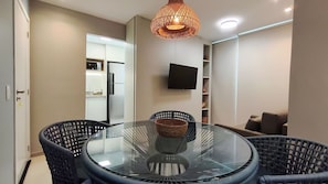 A modern dining area with a glass table, wicker chairs, a woven basket, a flat-screen TV, and a kitchen in the background.