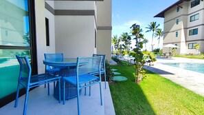 Un espace extérieur moderne aménagé avec une table en verre et des chaises bleues, un petit arbre, et une vue sur la piscine et un bâtiment bordé de palmiers.