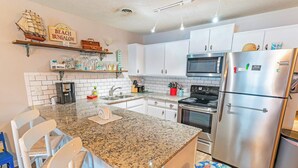 Una cocina con armarios blancos, un refrigerador de acero inoxidable, un microondas y una estufa. Hay taburetes de bar y una pequeña isla con encimera de granito.