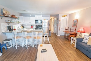 A compact kitchen with white cabinets, a microwave, and a refrigerator. There is a bar area with stools and a small table. The living area has a sofa and a lamp.