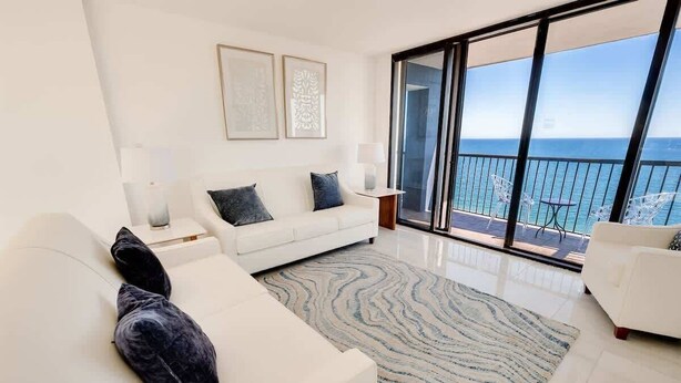 A modern living room with a white sofa, a patterned rug, and a balcony with a view of the sea.
