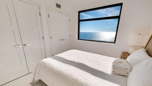 A bedroom with a bed, a window with a view of the sea, white walls, and a door.