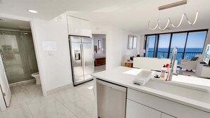 A modern kitchen with a large island, a refrigerator, and a view of the sea.