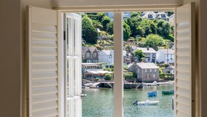 Una ventana con contraventanas abiertas que da a una masa de agua tranquila con barcos y edificios al fondo.