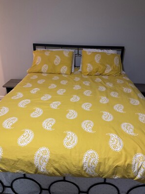 Ein Bett mit gelber Bettdecke mit weiĂen Paisleymustern, einem schwarzen Metall-KopfstĂŒck und einer weiĂen Wand im Hintergrund.