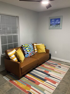 Uma sala de estar com um sofá marrom, almofadas coloridas, um tapete listrado, um ventilador de teto e um quadro na parede.