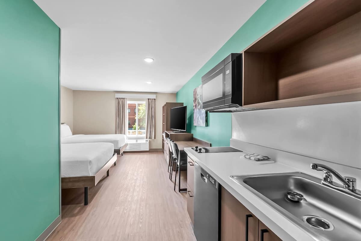 Quarto de hotel compacto com cozinha compacta, cama, televisão e janela com cortinas.
