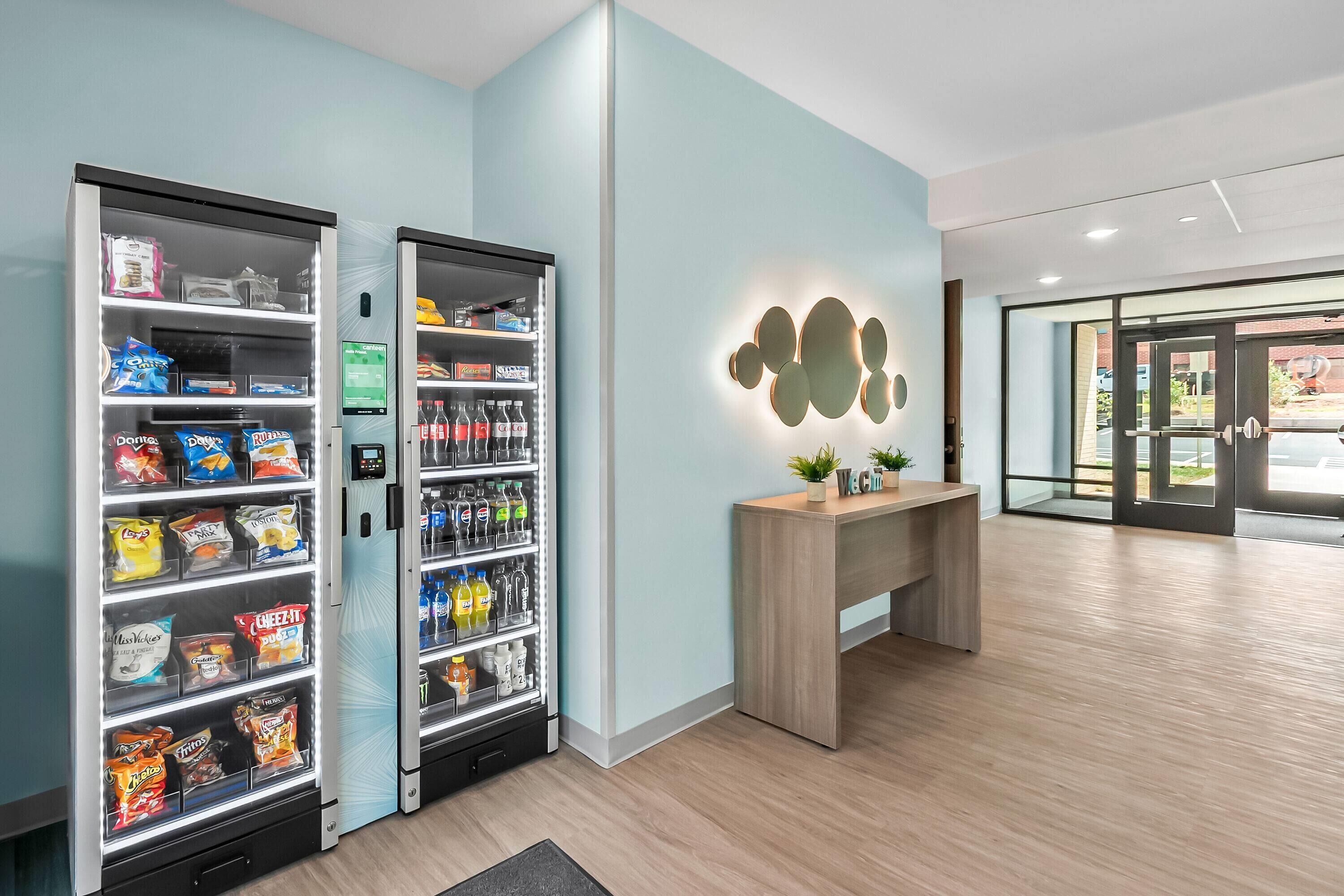 A clean, modern lobby with vending machines stocked with snacks and beverages, a wooden reception desk, and a minimalist wall design.