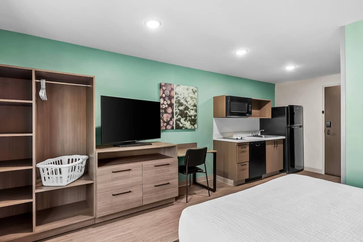 Quarto de hotel moderno com cama, televisão, secretária, cadeira e cozinha equipada com micro-ondas e frigorífico, além de uma cesto de roupa.