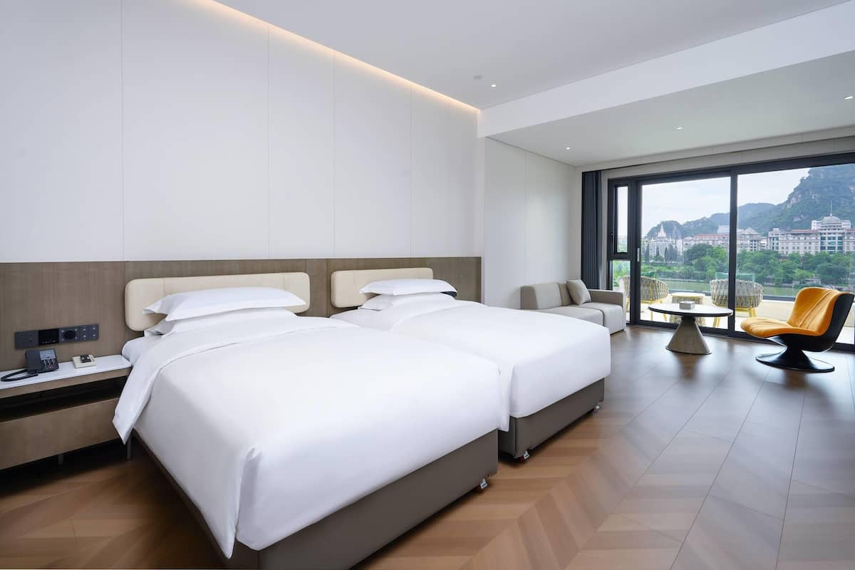 A modern hotel room with two beds, a sofa, a dining table, and a large window offering a city view.