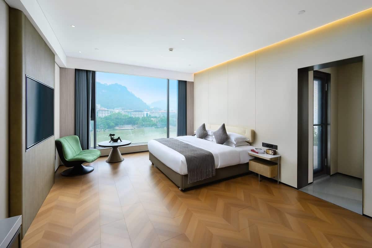 A modern hotel room with a large bed, a green chair, a small table, and a view of a city and mountains through large windows.