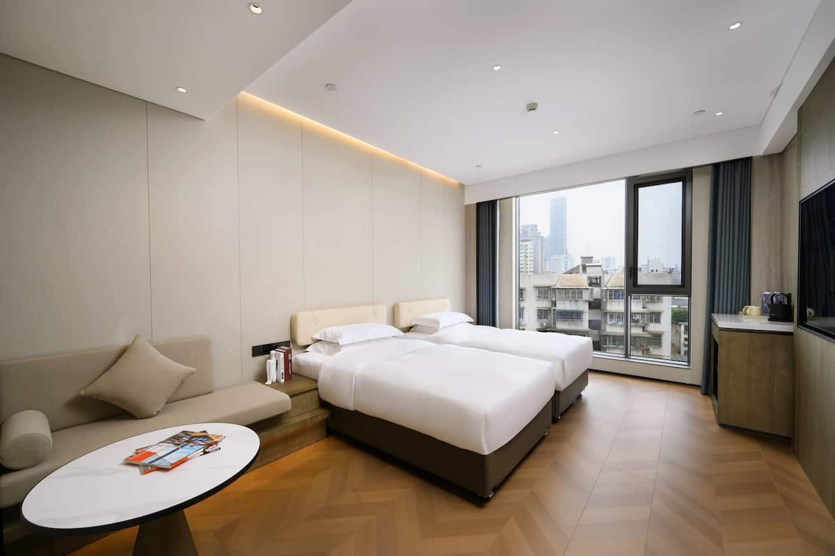 A modern hotel room with a sofa, two beds, a small table, and a large window offering a city view.