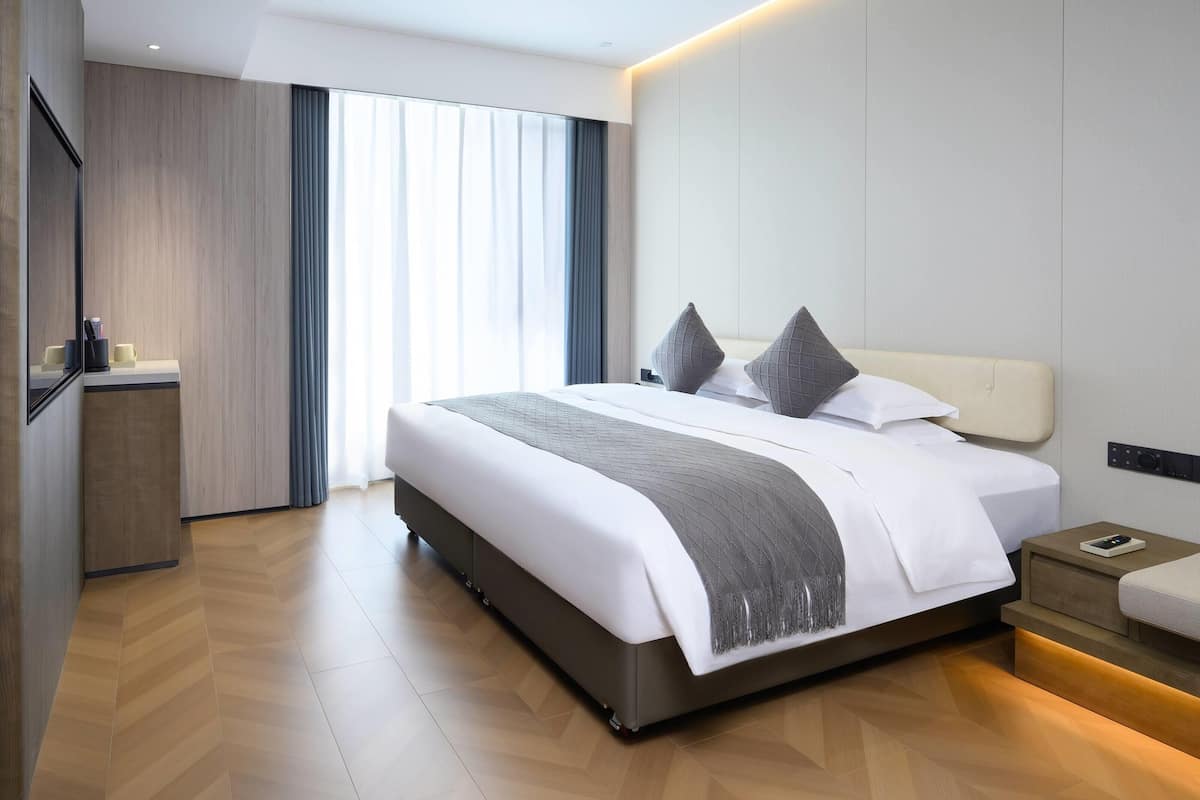 A modern hotel room with a large bed, bedside tables, a desk, and a large window with curtains.