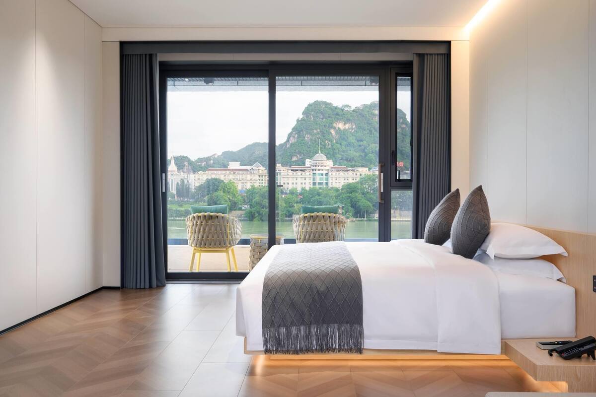 A modern hotel room with a large bed, a view of a cityscape and mountains, and a balcony with chairs.
