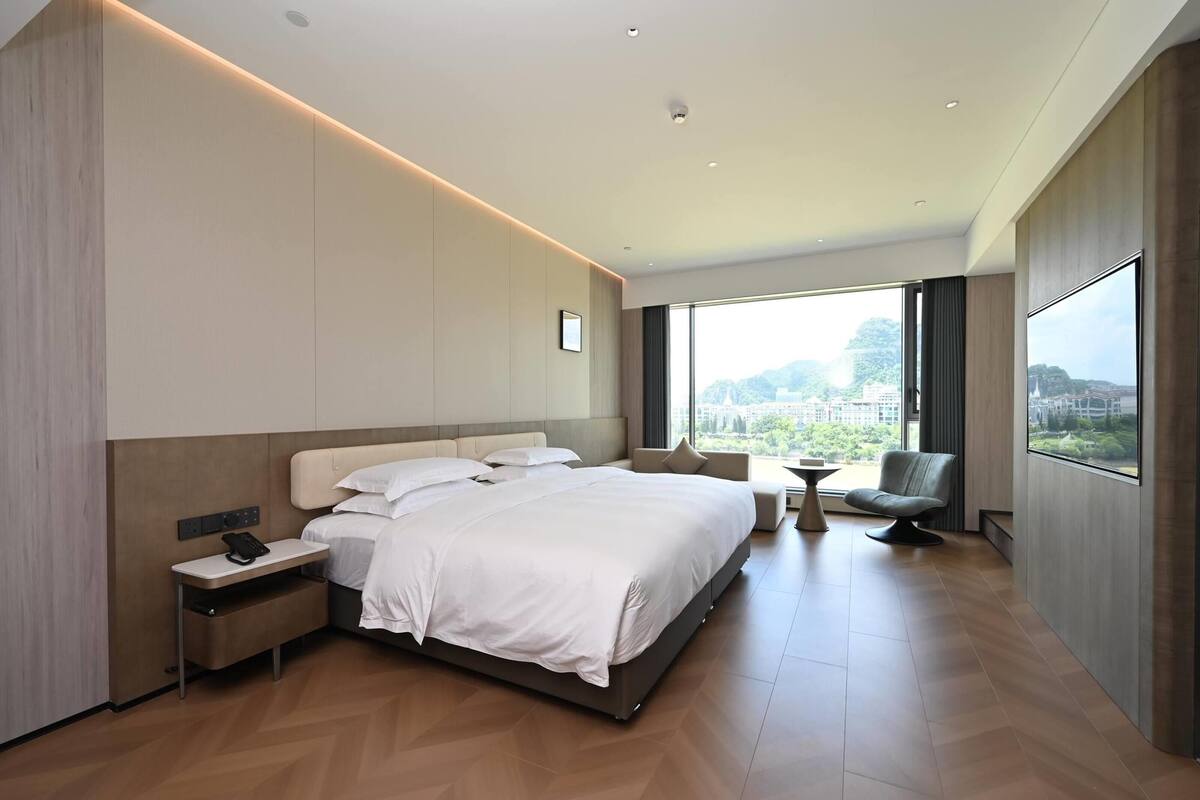A modern hotel room with a large bed, bedside tables, a seating area, and a view of the city.