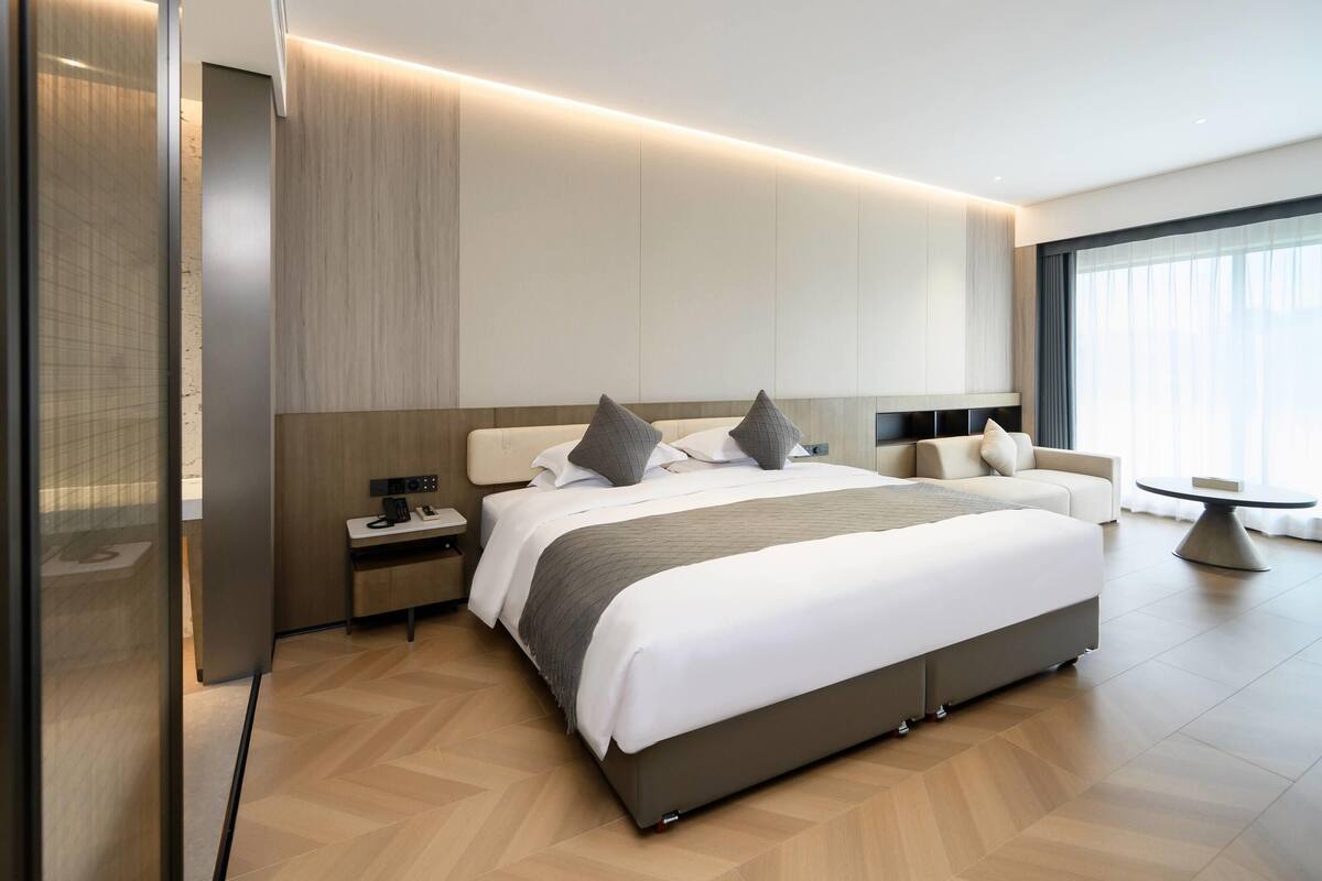 A modern hotel room with a large bed, bedside tables, a desk, and a view of the city through large windows.