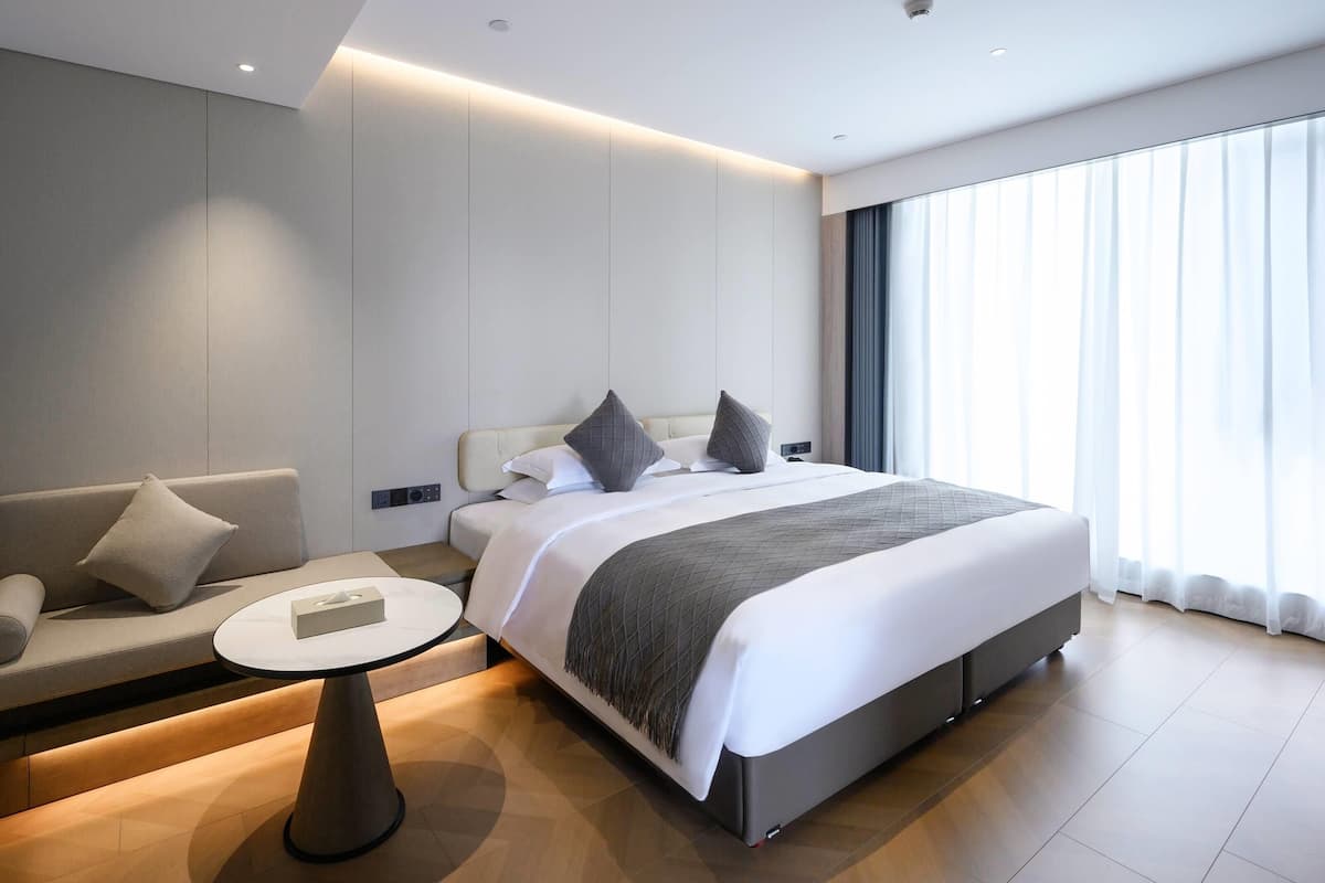 A modern hotel room with a large bed, a sofa, a small round table, and a large window with curtains.