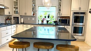 Una cucina moderna con isola centrale, tre sgabelli, mobili bianchi, elettrodomestici in acciaio inossidabile e una lampada a sospensione.