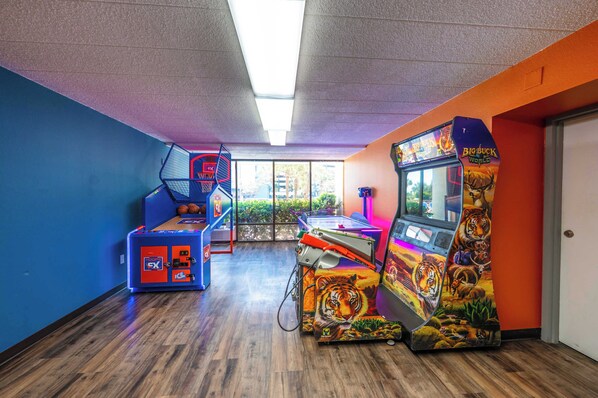 An indoor arcade with various gaming machines, a basketball hoop, and a window with a view of greenery.