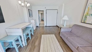 A modern living room with a sofa, a dining table with benches, a chandelier, and a door leading to another room. - New Direct Oceanfront, Pools Caravelle,HIGH Floor! (Myrtle Beach)