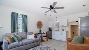 Ein Wohnzimmer mit grauem Sofa, einem braunen Ledersessel, Deckenventilator und einer Küchenzeile mit weißen Schränken und einem Edelstahlkühlschrank.
