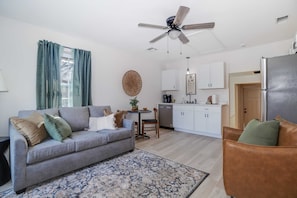 A living room with a grey sofa, a brown leather chair, a ceiling fan, and a kitchen area with white cabinets and a stainless steel refrigerator.