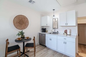 Una cocina compacta con armarios blancos, lavavajillas, cafetera y una pequeña mesa de comedor con sillas.