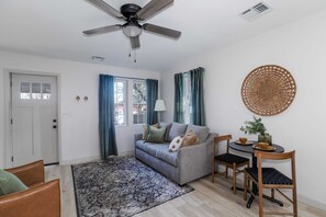 Una sala de estar con sofá, sillas, una mesa redonda con una planta, un ventilador de techo y una puerta.