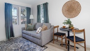 Ein Wohnzimmer mit grauem Sofa, zwei Holzstühlen, einem runden Tisch mit gewebter Tischdecke und einem blau gemusterten Teppich.
